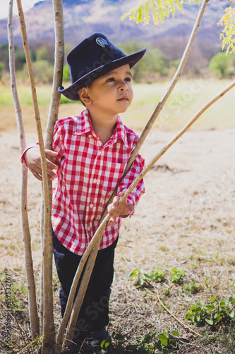 Child dressed as a cowboy leaning against a tree. White boy in cowboy clothes outdoors. Child smiling. Having fun in the park. Child in contact with nature. Child looking to the side. 