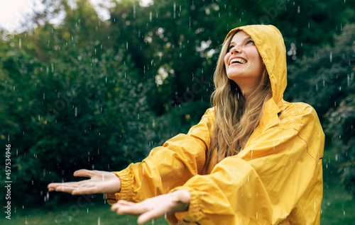 Horizontal side view image of a joyful young woman smiling wearing yellow raincoat during the rain in the nature. Pretty female looking up and catching the rain drop with hands outdoors in the park. photo