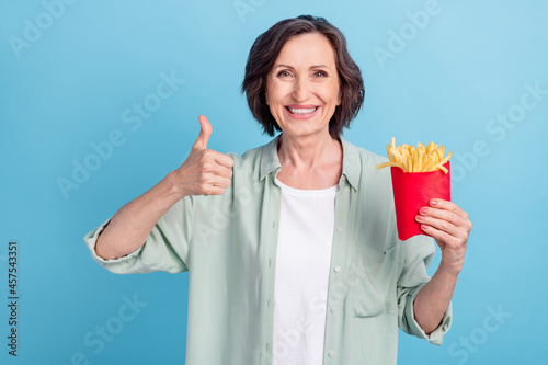 Photo portrait senior woman in shirt eating fried potatoes showing thumb-up isolated pastel blue color background photo