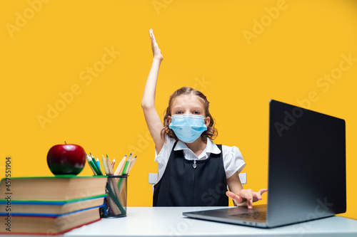  Schoolgirl raising hand up and sitting at laptop wearing mask. Yellow background. Distance Learning photo