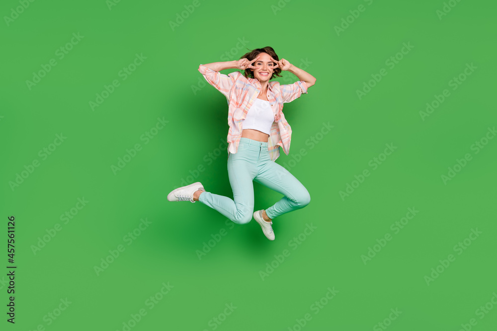 Full length photo of cheerful happy young woman jump up hands v-signs face enjoy isolated on green color background