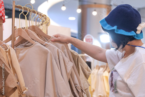 Woman wears beanie with protective mask choosing neutral cotton clothes on clothing rack in fashion store at shopping mall