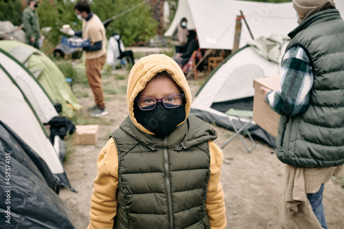 Portrait of mixed race child in hoodie, vest, eyeglasses and cloth mask standing in refugee camp © pressmaster
