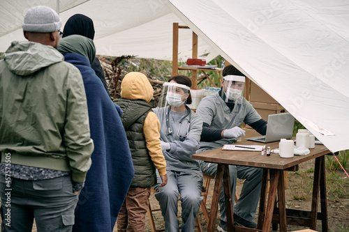 Medical specialists in protective masks sitting at table in tent and checking health of migrants standing in line