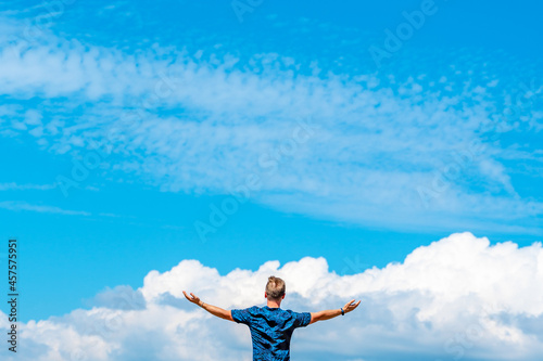 Rear view of a young man standing with his arms open against the background of a blue sky with clouds. The concept of freedom