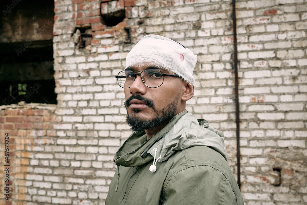 Portrait of miserable young Middle-eastern man with beard having bandaged head, abandoned building in background Portrait of miserable young Middle-eastern man with beard having bandaged head, abandoned building in background