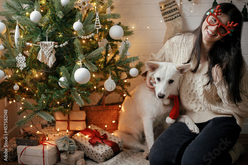 Stylish happy woman and adorable dog sitting under christmas tree with gifts and lights. Young female and cute white dog in festive accessories celebrating in scandinavian room. Happy Holidays!