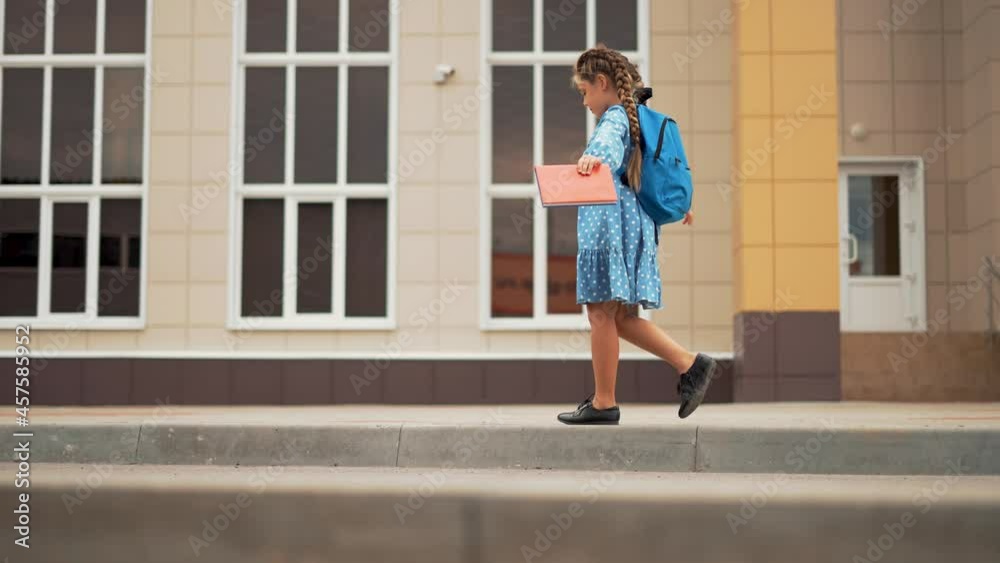 Happy girl goes with backpack to school. Girl holding book in her hands goes back to school. Girl is first to school. Happy girl back to school. Child with backpack in schoolyard. Schoolgirl with book