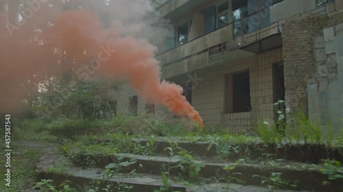 Hand flare emitting orange smoke against background of old building in abandoned forest area. Dilapidated concrete stairs overgrown with grass on foreground photo