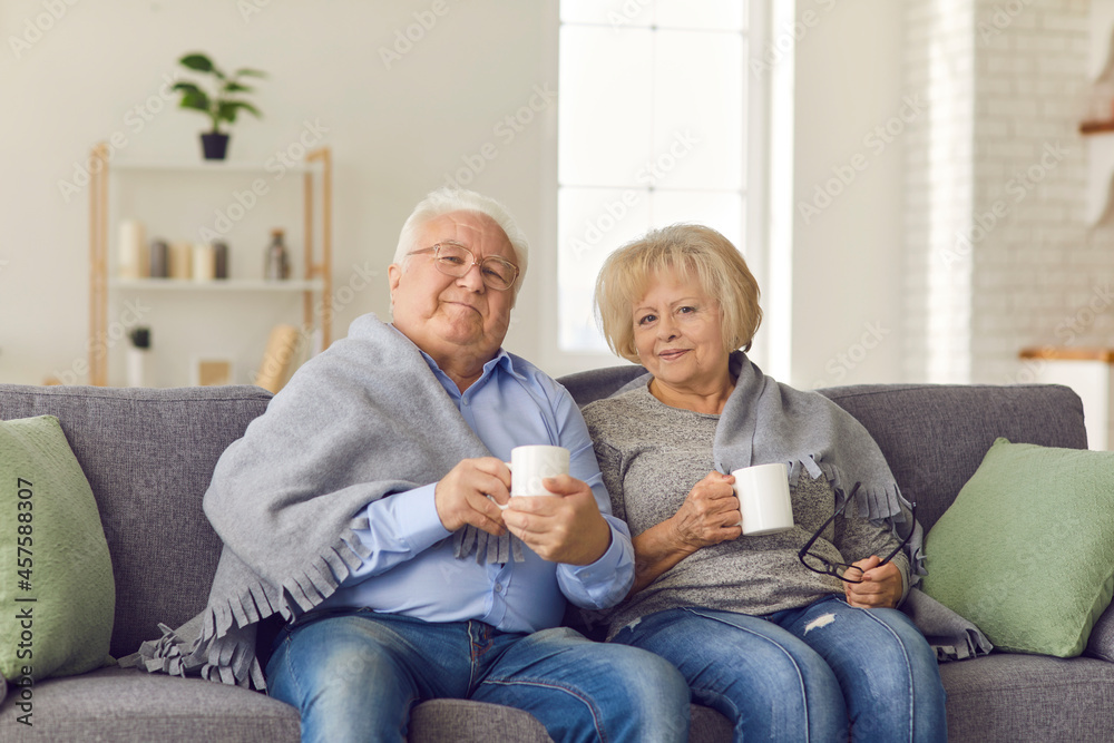 Happy mature couple sitting together on sofa under blanket and holding hot drinks in cups in hands at home with room interior at background. Elderly people happy lifestyle, family happiness concept