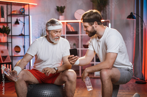 Middle aged man showing something on smartphone to pensioner that sitting on fitball. Two caucasian persons wearing sport clothes at home. Evening workout of son and father.
