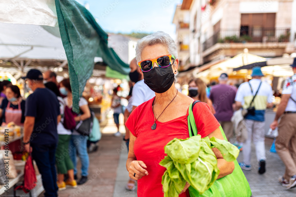 Old lesbian woman in protective medical mask and sunglasses with lgbt pride flag buying food at a market on the city street.