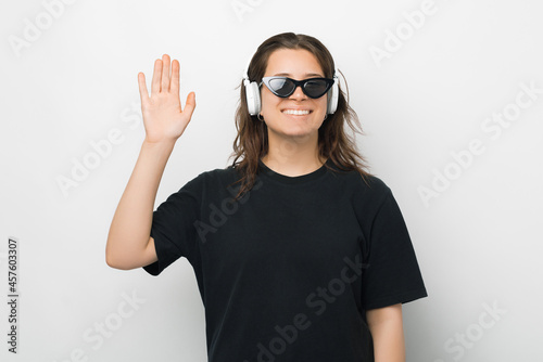 Relaxed girl listening to the music through headphones is waving at the camera over white background.