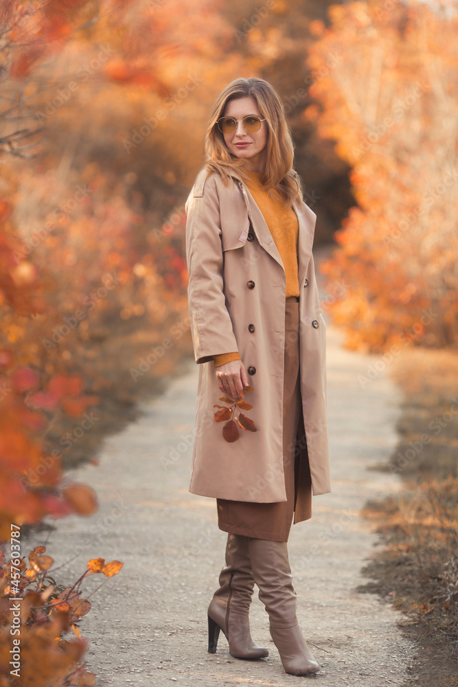 Fashionable and beautiful smiling young woman 30-35 years old, dressed in an orange knitted sweater, yellow glasses, brown wrap skirt and a beige classic trench coat. Fashion clothes autumn look. Fashionable and beautiful smiling young woman 30-35 years old, dressed in an orange knitted sweater, yellow glasses, brown wrap skirt and a beige classic trench coat. Fashion clothes autumn look.