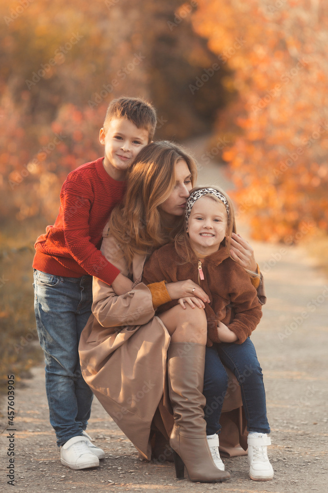 Fototapeta premium Portrait of a caucasian mother hugging and kissing her children in beautiful outfits on a sunny autumn day in nature park. Family happiness concept.