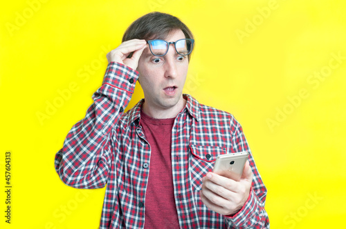 handsome excited man with open mouth in red checkered shirt taking off glasses on forehead and looking at smartphone on yellow background with copy space
