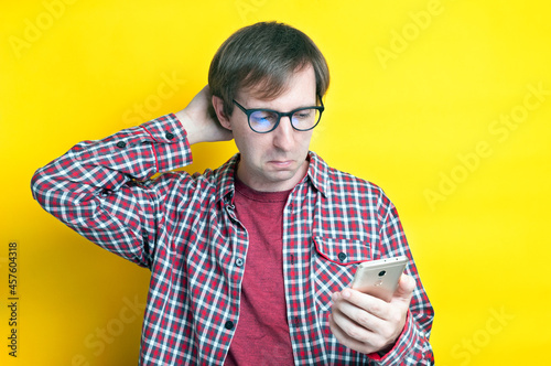 handsome confused man in checkered shirt with hand on back of head looking at smartphone on yellow background with copy space