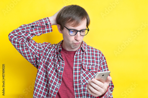 handsome upset man in checkered shirt with hand on back of head and in glasses looking at smartphone on yellow background with copy space