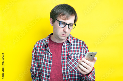 handsome sad man in red checkered shirt and glasses looking at smartphone on yellow background with copy space