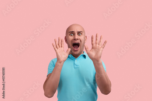 Frightened bald homosexual man with bristle, screams in fright, hold hands near face in protective gesture, has scared expression, gay friendly, wears blue polo shirt, poses over pink background.