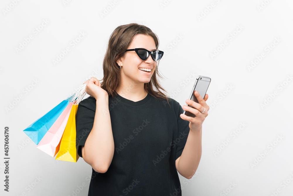 Smiling girl wearing sunglasses is ordering something online by her phone over white background.