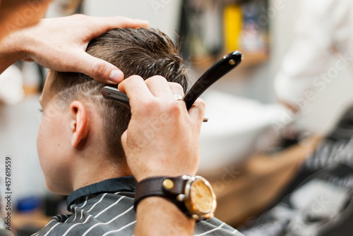 Male hands hold a straight razor near child's head. Men's haircut in a barbershop. Male hairdresser cuts boy's hair during the COVID-19 epidemic. Accessories for haircuts and hair care. Beauty saloon © Sofiia