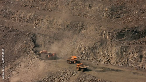 Two heavy dump trucks drive up to an excavator loading iron ore in a quarry time lapse.Machines are removing ore out of the mine site. Iindustrial tourism concept. Open pit vehicles work in the mine.  photo