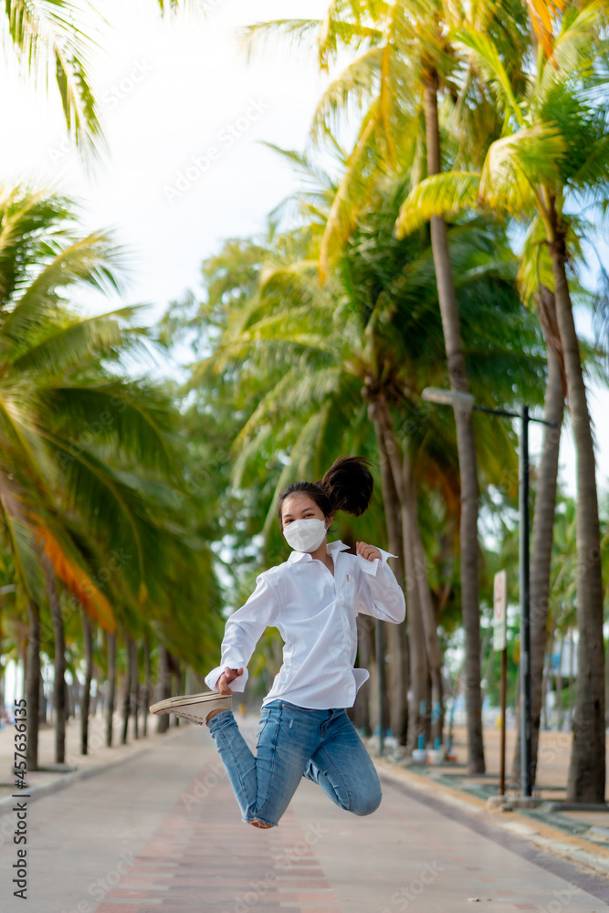 A young Asian woman wearing a white shirt and jeans is posing for a photo on the Bangsaen beach walkway on a day when there are not many people behind the coconut trees. Bangsaen, Thailand