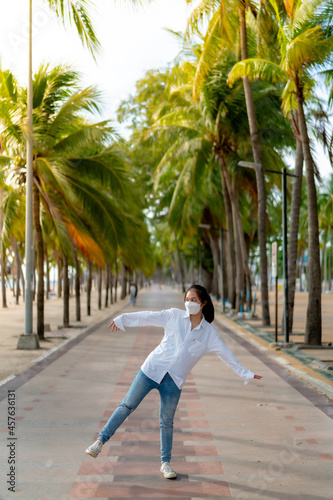 A young Asian woman wearing a white shirt and jeans is posing for a photo on the Bangsaen beach walkway on a day when there are not many people behind the coconut trees. Bangsaen  Thailand