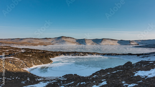 Fototapeta Naklejka Na Ścianę i Meble -  A frozen lake in the form of an ice heart. Snow lies on the ground, devoid of vegetation.  Nearby is Lake Baikal. A mountain range against the blue sky. Siberia