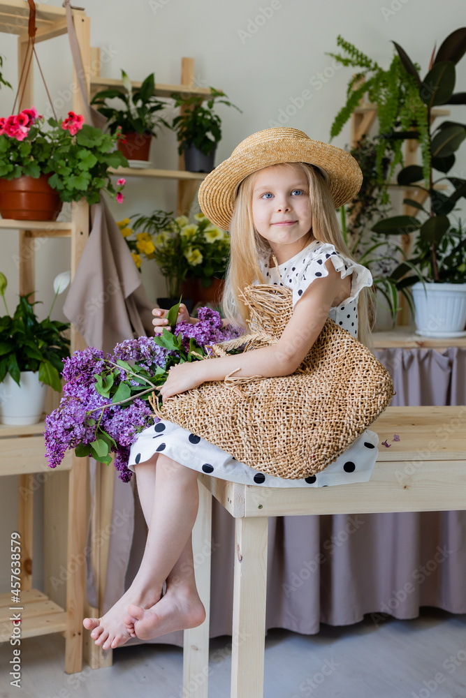 Fototapeta premium a little blonde girl in a dress with black polka dots and a straw hat with a bouquet of lilac flowers is sitting on a wooden chair in the room. lifestyle, high quality photo