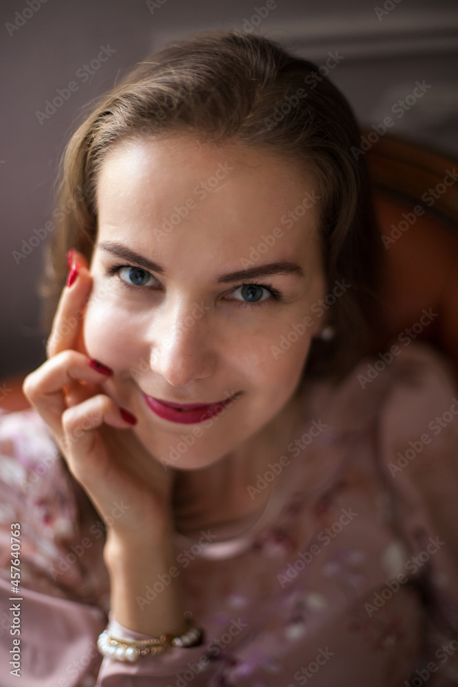 Young beautiful woman in pink dress and stylish accessories sits on brown leather sofa on white wall background. Portrait of happy smiling girl with red lips resting at home and looking at camera. Young beautiful woman in pink dress and stylish accessories sits on brown leather sofa on white wall background. Portrait of happy smiling girl with red lips resting at home and looking at camera.