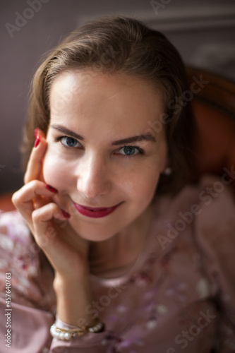 Young beautiful woman in pink dress and stylish accessories sits on brown leather sofa on white wall background. Portrait of happy smiling girl with red lips resting at home and looking at camera. 