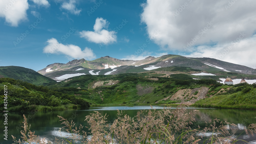 The emerald water of the lake is calm.  A mountain range against a background of blue sky and clouds. There are areas of snow on the slopes. In the foreground - spikelets of grass. Kamchatka