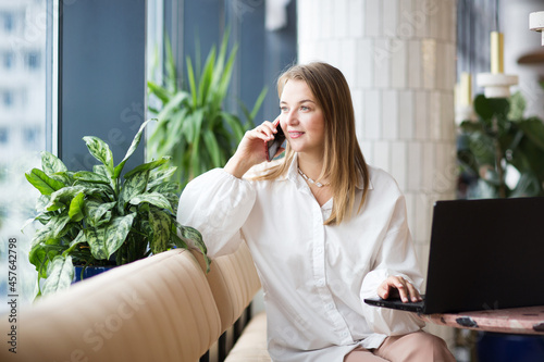 successful business woman sitting in cafe with laptop, working and talking on phone. office workplace with green plants.