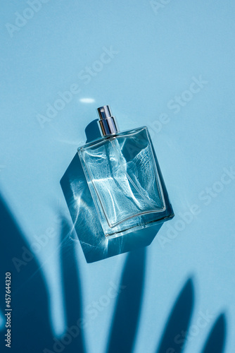 Transparent bottle of perfume on a blue background. Fragrance presentation with daylight. Trending concept in natural materials with palm leaves shadow. Women's and men's essence.