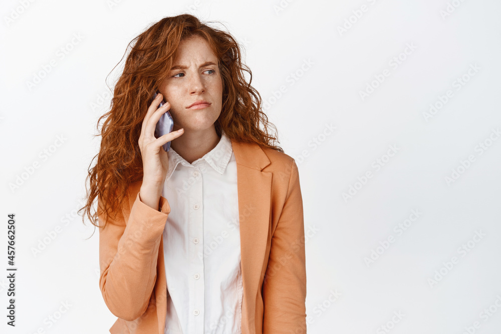 Annoyed redhead girl calling someone, waiting for an answer on smartphone, frowning and grimacing, standing over white background Annoyed redhead girl calling someone, waiting for an answer on smartphone, frowning and grimacing, standing over white background