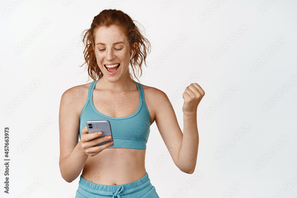 Happy sportswoman looking at her fitness app on mobile phone and triumphing, achieve daily goal in sport app, standing in activewear against white background Happy sportswoman looking at her fitness app on mobile phone and triumphing, achieve daily goal in sport app, standing in activewear against white background