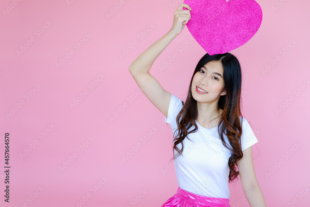 Portrait beautiful asian teen girl wearing white T-shirt and pink skirt on pink background, happy valentine day in love concept, model holding red heart sign in hand Portrait beautiful asian teen girl wearing white T-shirt and pink skirt on pink background, happy valentine day in love concept, model holding red heart sign in hand