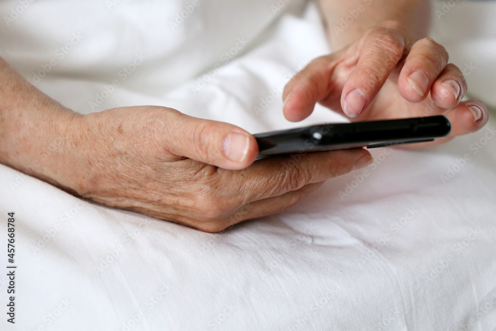 Elderly woman with smartphone in a bed, mobile phone in wrinkled female hands close up. Concept of online communication in retirement, sms, social media Elderly woman with smartphone in a bed, mobile phone in wrinkled female hands close up. Concept of online communication in retirement, sms, social media