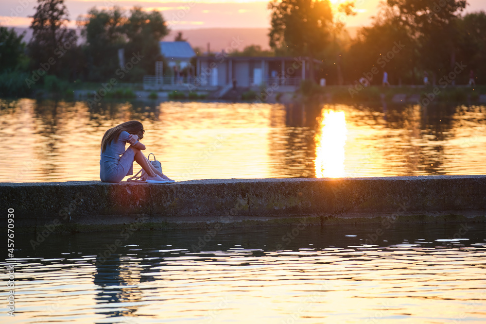 Side view of lonely woman sitting alone on lake shore on warm evening. Solitude and relaxing in nature concept.