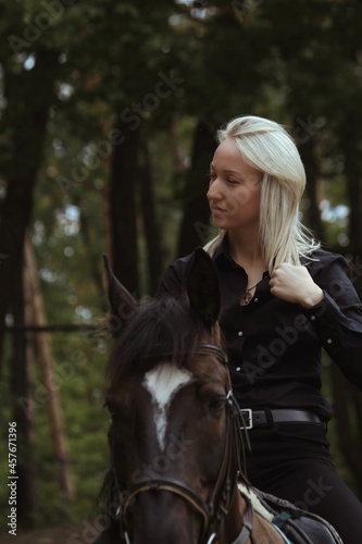 A blonde girl in profile riding a black horse against a green forest. A girl is riding a brown horse on a autumn day.