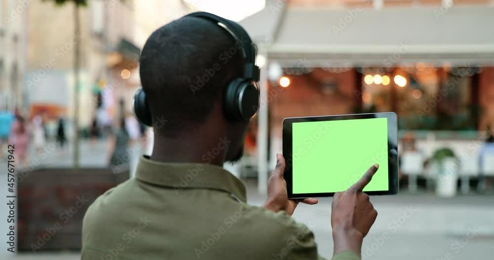 Close up of the multiracial male wearing headphones holding a horizontal black tablet with green empty screen in the middle of the city street. Chroma key concept