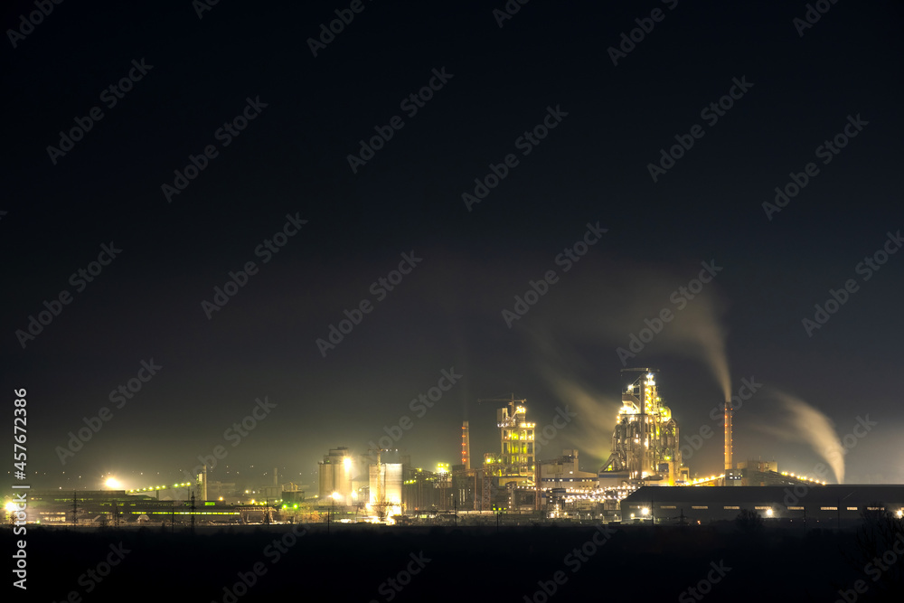 High concrete structure of cement factory with tower crane and smoking chimney at night. Industrial production and air pollution concept.