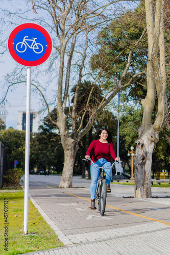 young hispanic latina woman riding her bicycle smiling on the bike path. bike traffic sign. in the city in autumn. vertical photo