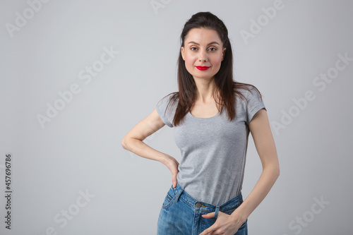 Portrait of pretty young brunette woman in mid 30s wearing casual clothes. Cute female model in grey t-shirt and classic blue jeans on grey background in studio