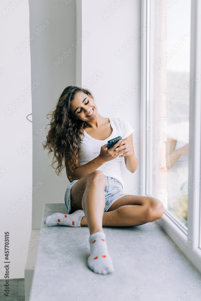 Young woman sitting on the windowsill and writing message on smartphone at home