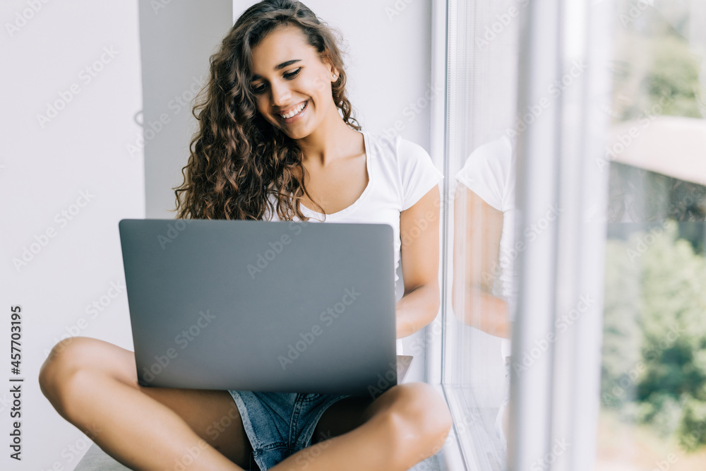 Young pretty woman sitting near the window with a laptop, she uses the computer at home on the windowsill Young pretty woman sitting near the window with a laptop, she uses the computer at home on the windowsill