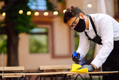 Disinfecting to prevent COVID-19. Waiter cleaning the table with Disinfectant Spray in a restaurant wearing protective medical mask and gloves.