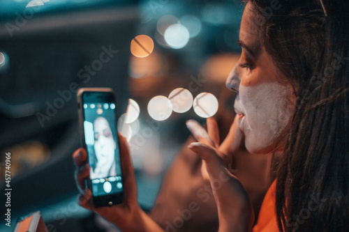 Adolescente joven pintándose por la noche mientras se observa en el teléfono móvil en un coche sentada con iluminación de la ciudad photo
